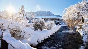 A snow-covered landscape with a building, a flowing river, and snow-laden trees. - Spa & Resort Bachmair Weissach, LUXURY FAMILY RESORT (Kreuth)