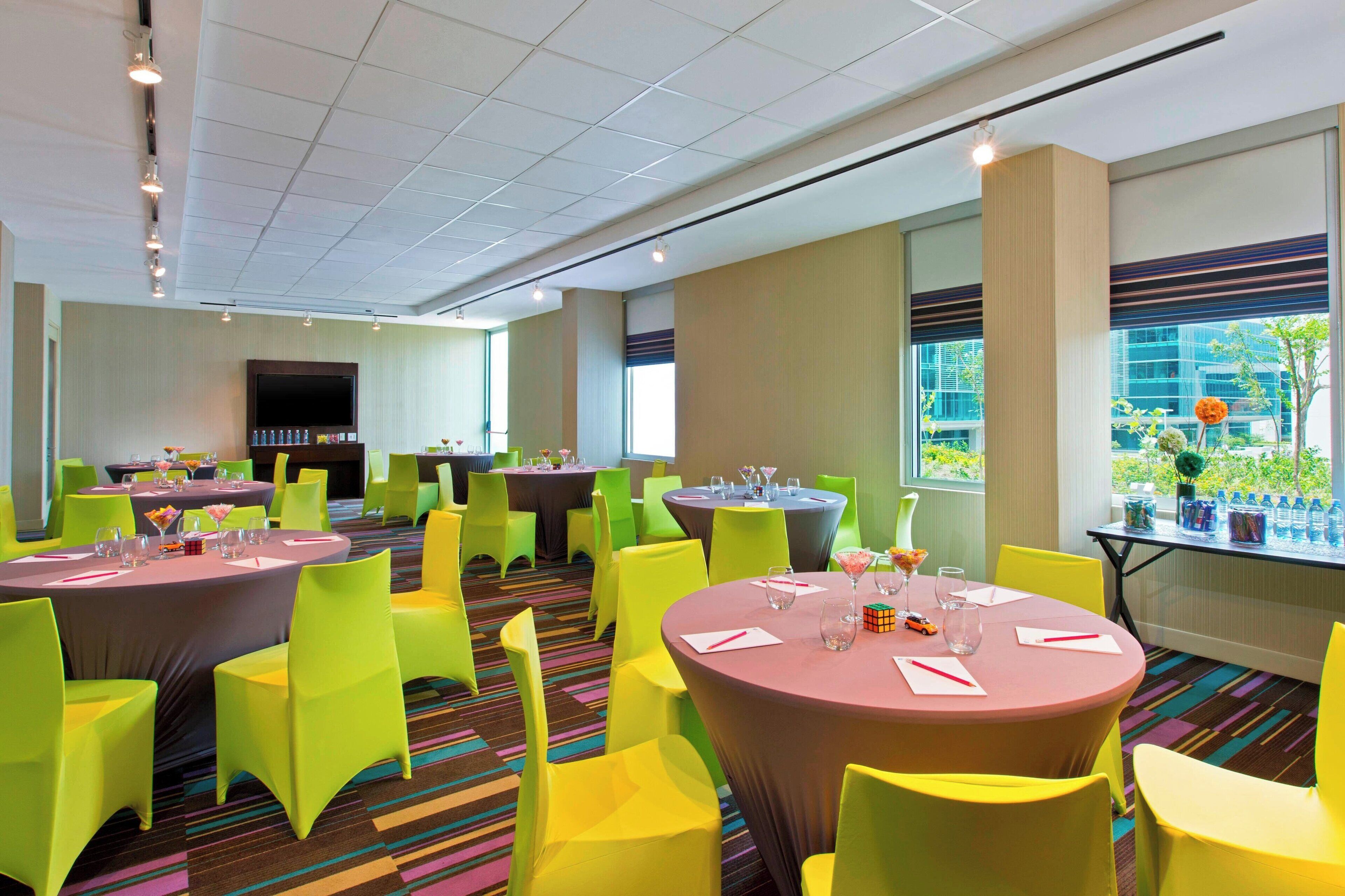A hotel dining area with multiple round tables, bright yellow chairs, and a large window with a view of greenery.