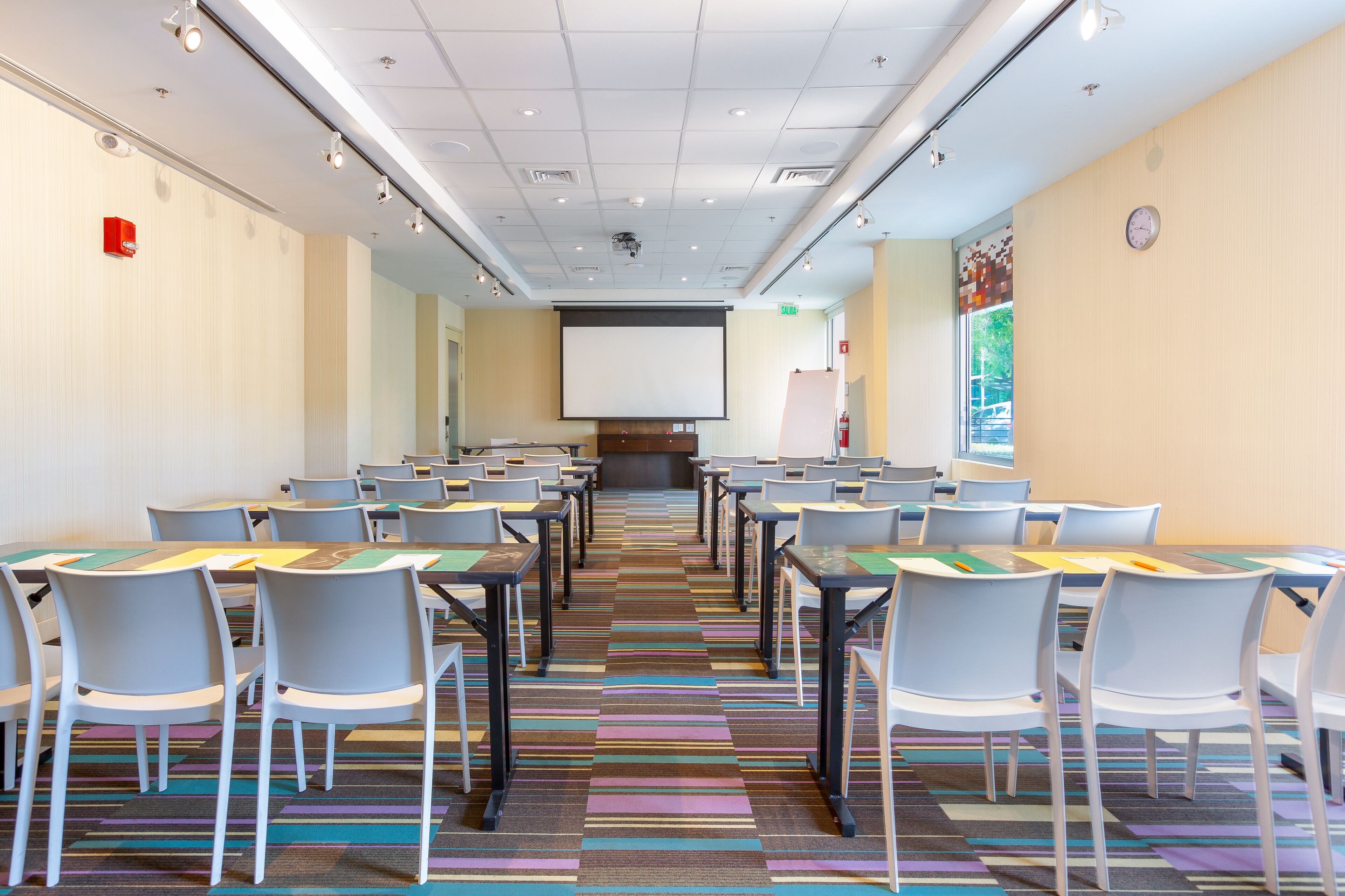 A conference room with rows of tables and chairs, a projector screen, and a patterned carpet.