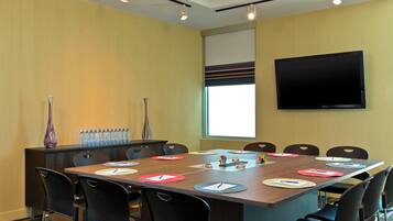 A conference room with a long table, black chairs, a TV on the wall, and a window with blinds.