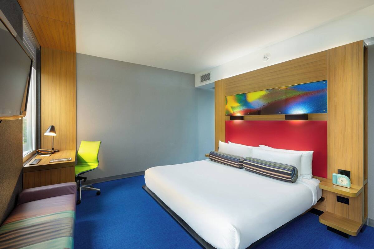 A hotel room with a large bed, a desk with a chair, a window, and a colorful artwork on the wall.