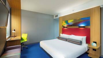 A hotel room with a large bed, a desk with a chair, a window, and a colorful artwork on the wall.