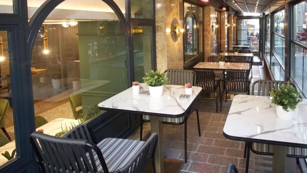 Un comedor interior con mesas y sillas de mármol, plantas en macetas y ventanas amplias con vistas a la calle.
