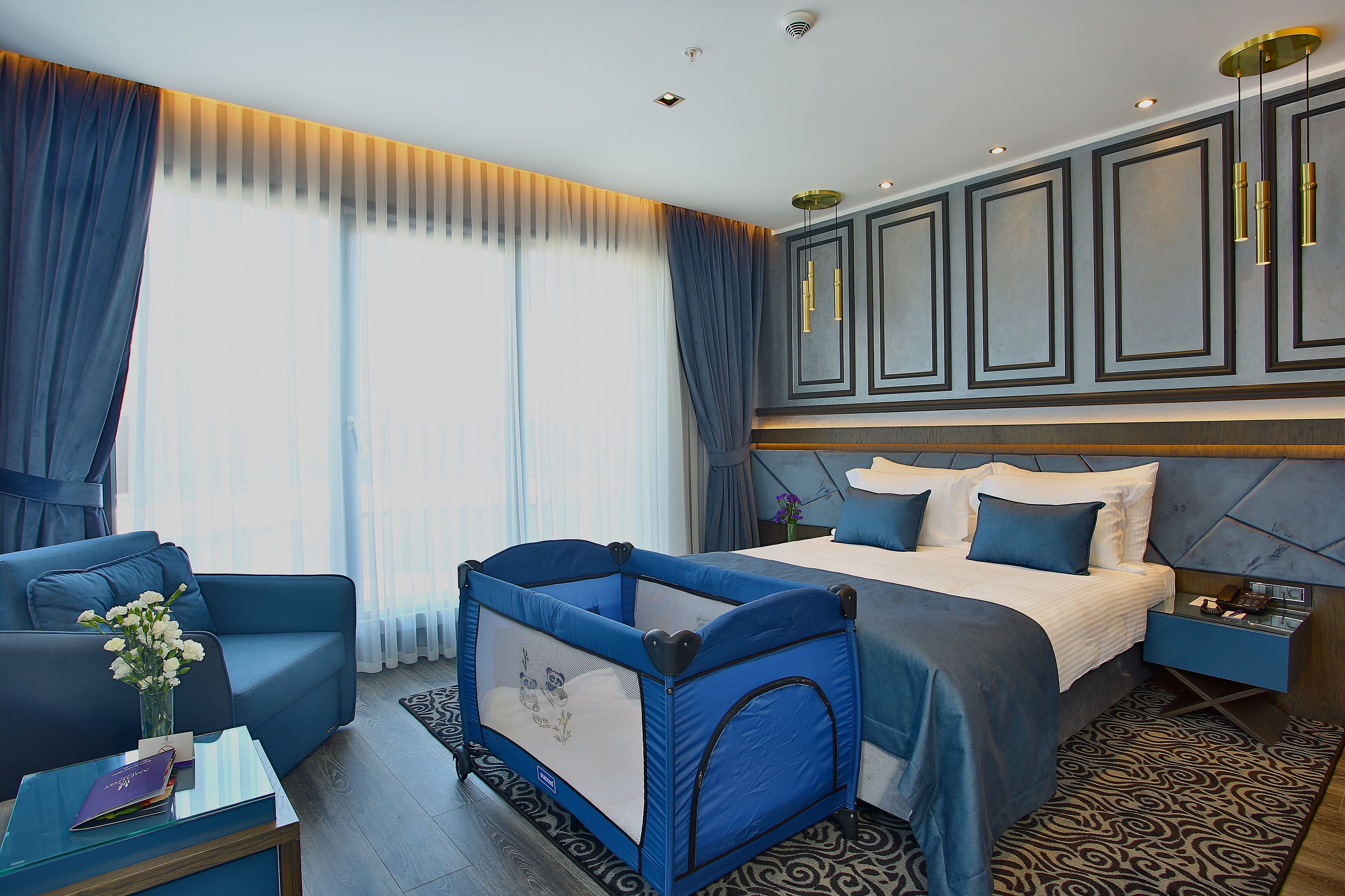 A hotel room with a large bed, a blue sofa, a small playpen, and a nightstand with a lamp.