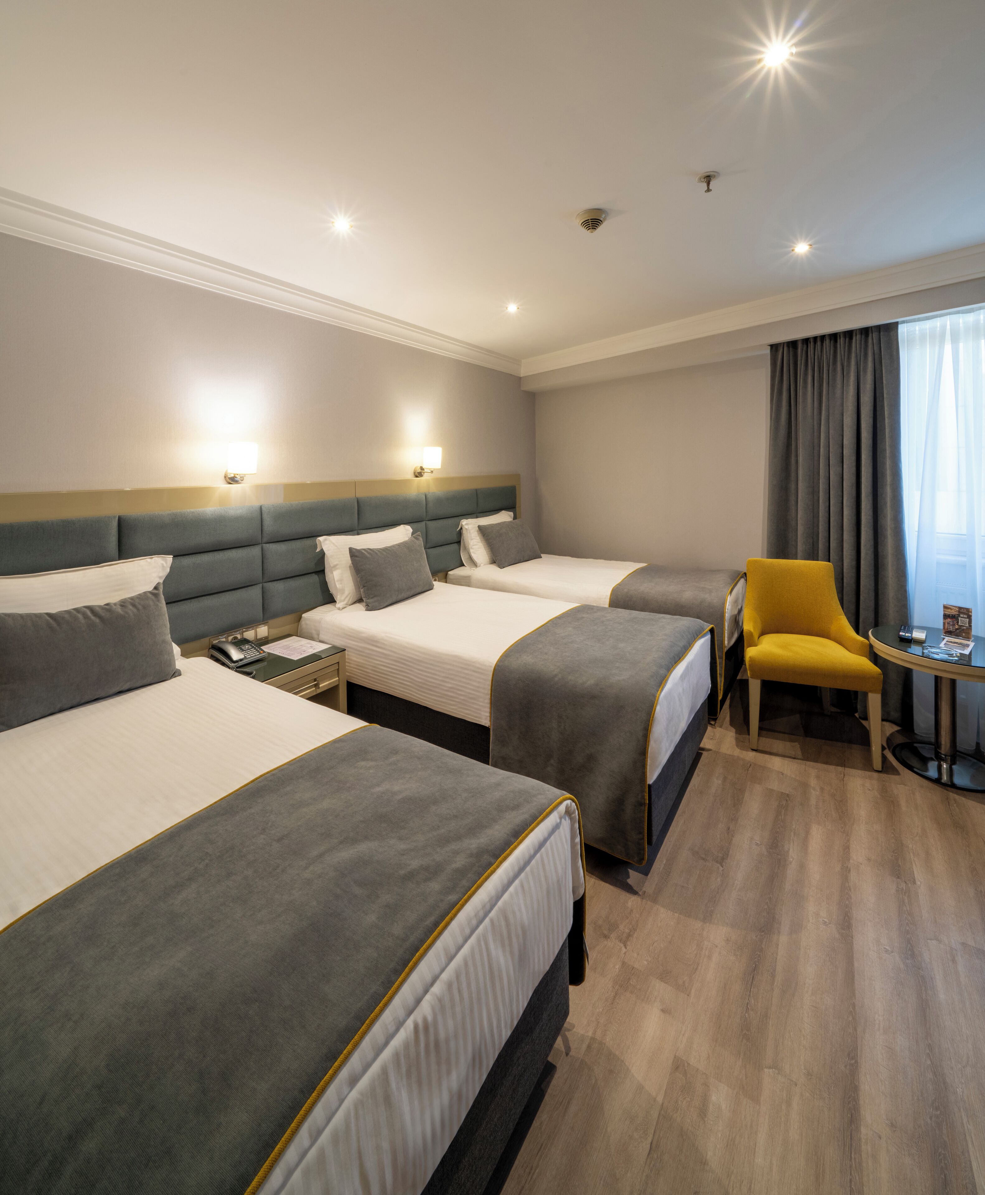 A hotel room with three beds, a small table with a chair, and a window with curtains.