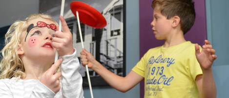 Two children playing with frisbees indoors, one wearing a white shirt with a pink design and the other in a yellow shirt with text.