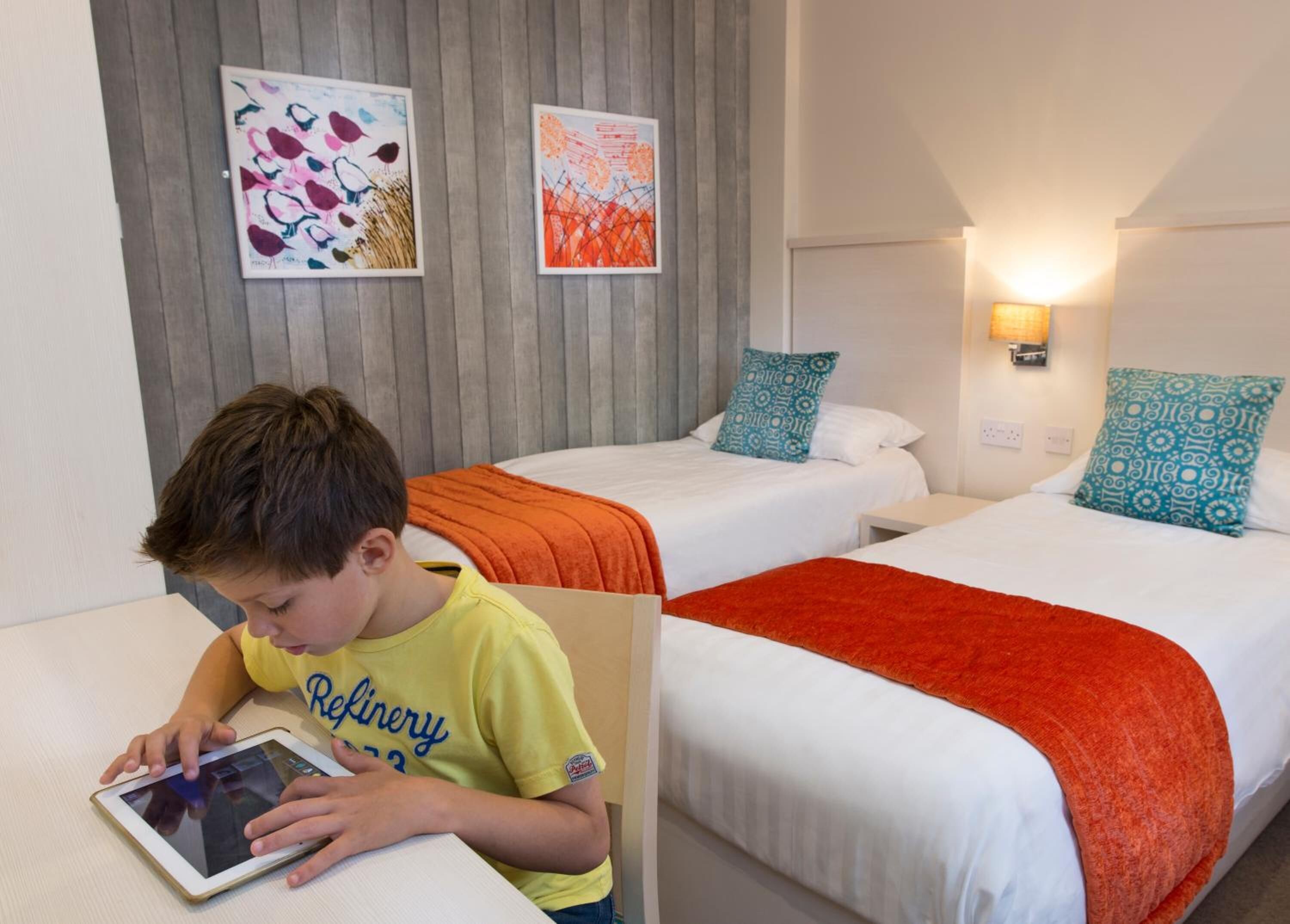 Hotellihuone, jossa on kaksi sänkyä, lapsi käyttää tablettia, ja seinillä on kaksi taideteosta.