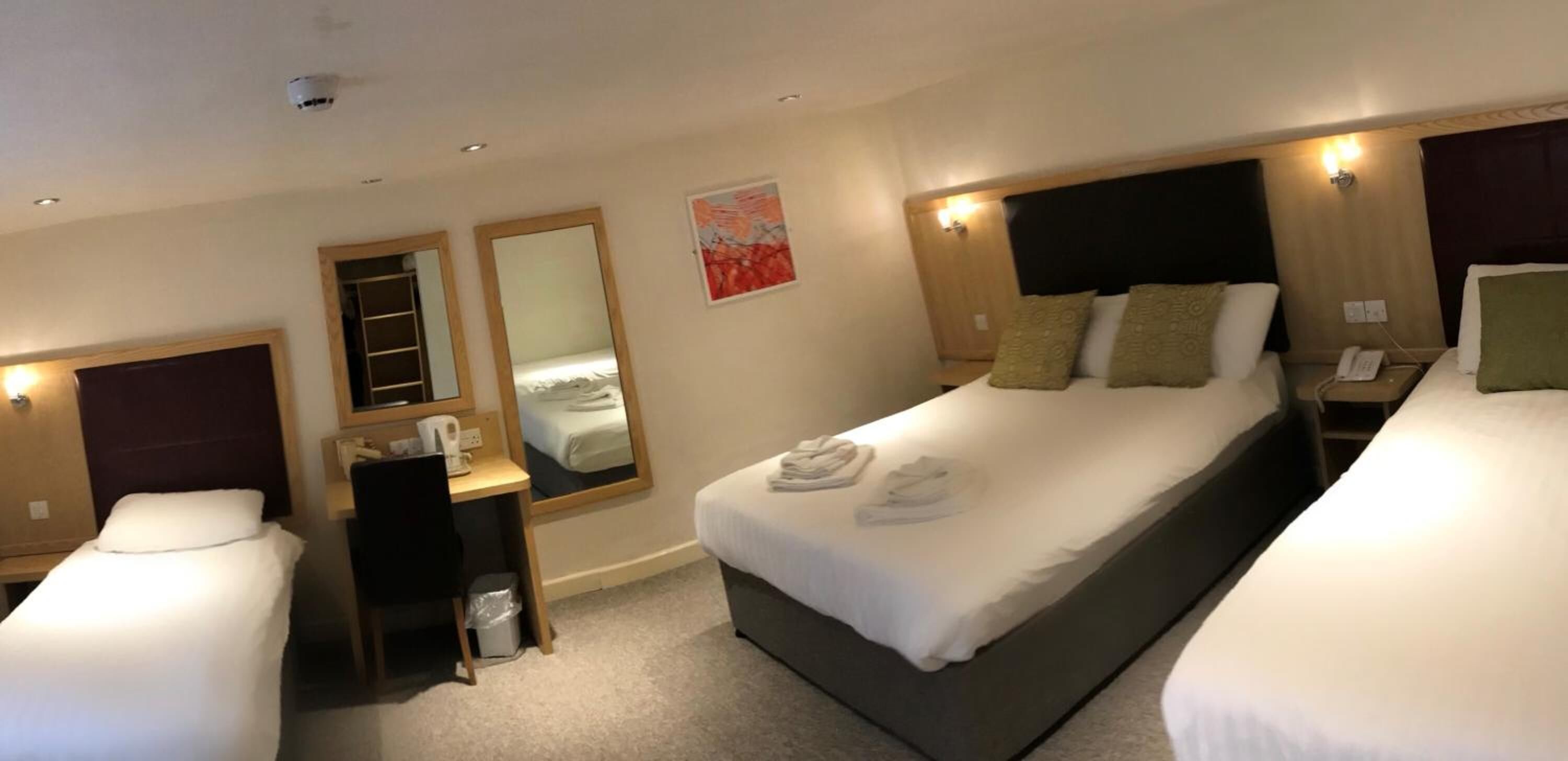 Quarto de hotel com duas camas de solteiro, uma escrivaninha com cadeira, um espelho e uma pequena lixeira.