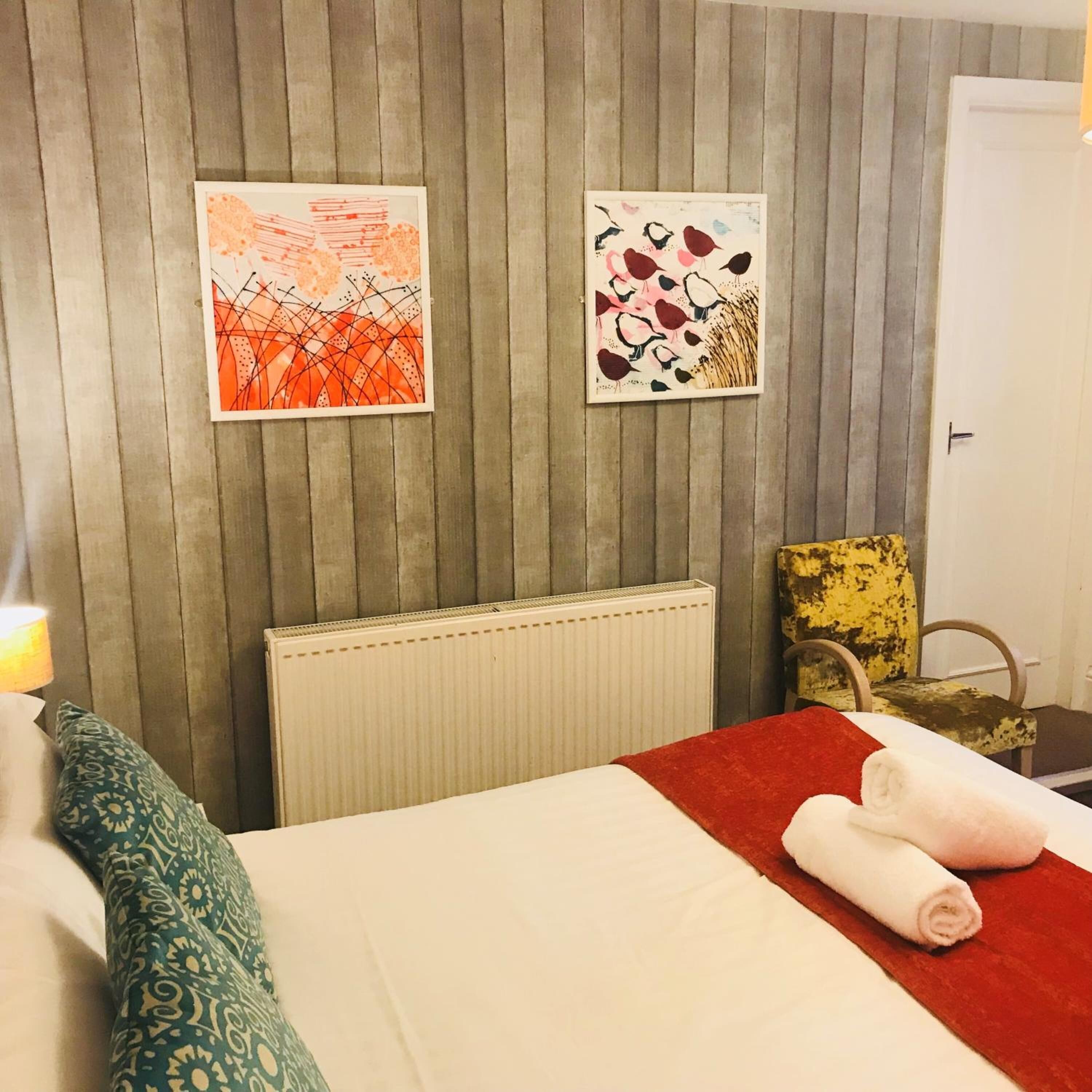 Ein Hotelzimmer mit Bett, Heizkörper, zwei Kunstwerken an der Wand und einer roten Tagesdecke.