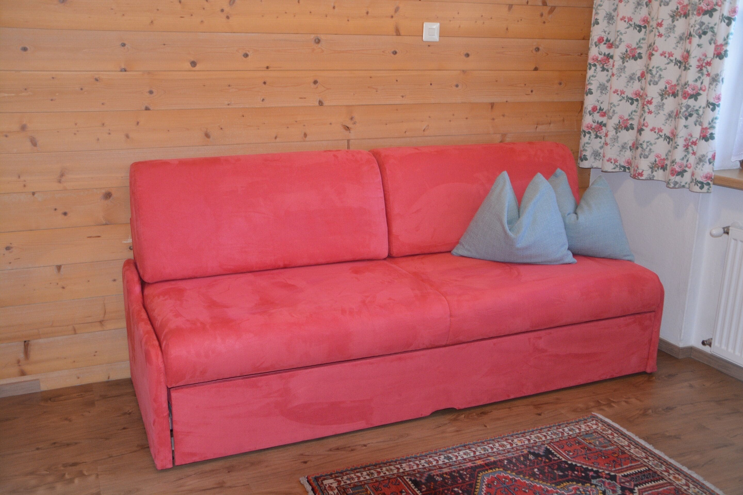 Un canapé rouge avec des coussins gris, adossé à un mur en bois, un tapis à motifs sur le sol et une fenêtre ornée de rideaux à fleurs.