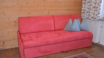 Un sofá rojo con cojines grises frente a una pared de madera, una alfombra estampada en el suelo y una ventana con cortinas florales.