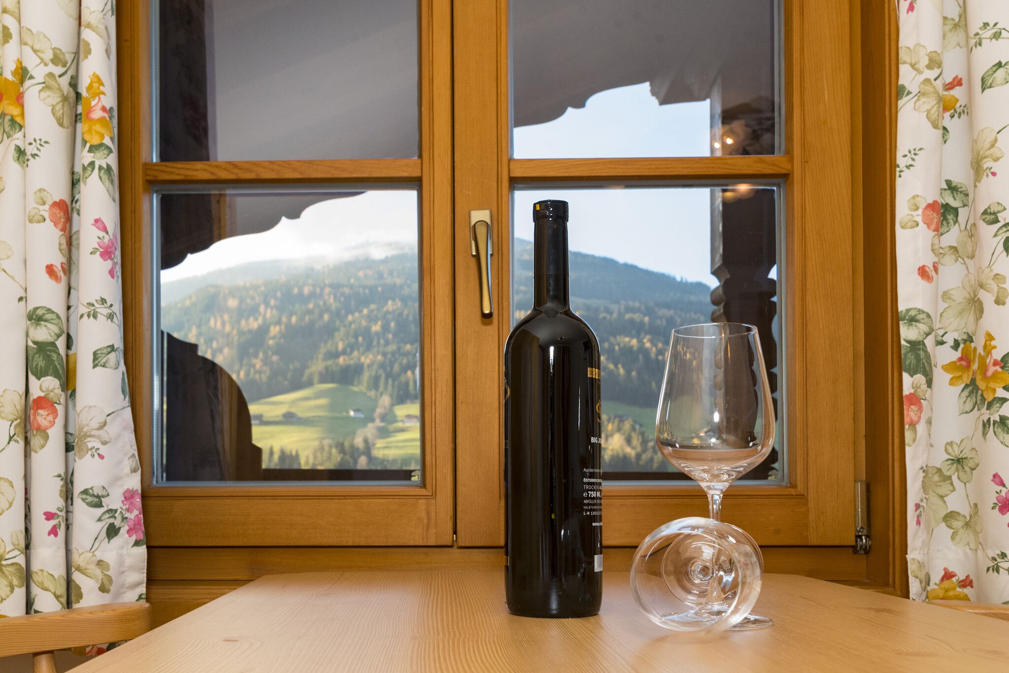Eine Weinflasche und ein Weinglas auf einem Tisch vor einem Fenster mit Blick auf eine gebirgige Landschaft.