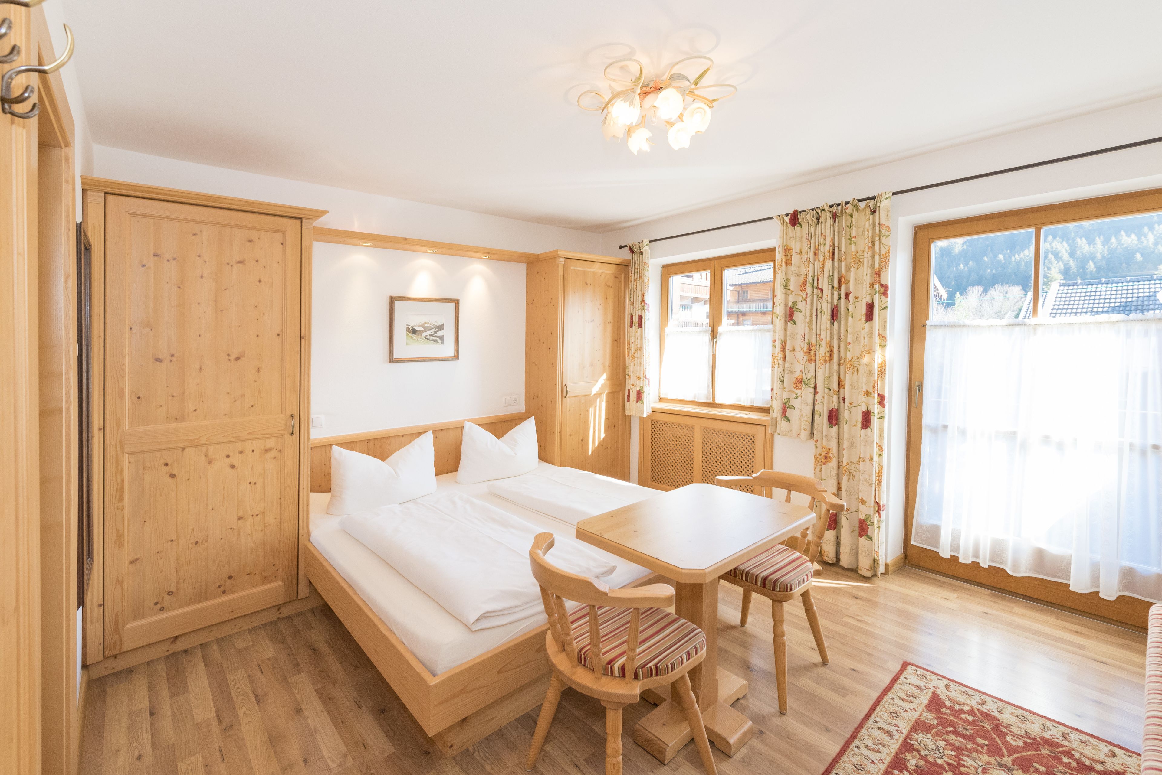 Una habitación con una cama de madera, una mesa de comedor, sillas, un tapete pequeño y una ventana con cortinas.