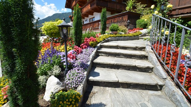 Una escalera de piedra conduce a un edificio de madera con balcones, rodeado de macizos de flores coloridos y vegetación.