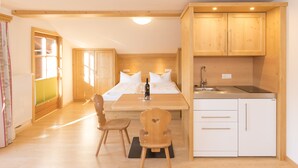 Una habitación pequeña y acogedora con cama, una mesa de madera con sillas, una zona de cocina con lavaplatos y armarios, y una ventana con cortinas.