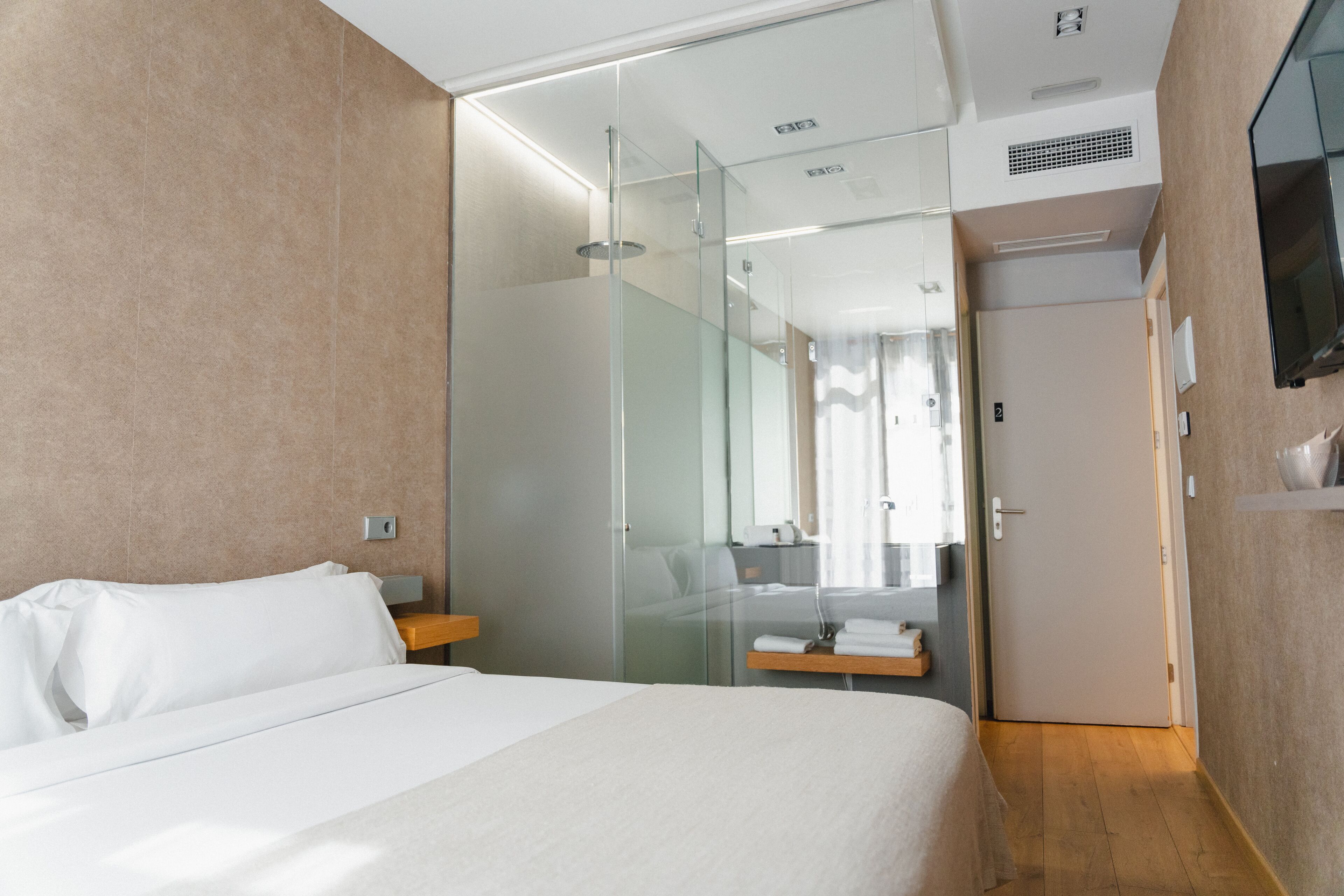 Habitación de hotel moderna con una cama grande, cabina de ducha de vidrio y un televisor montado en la pared.