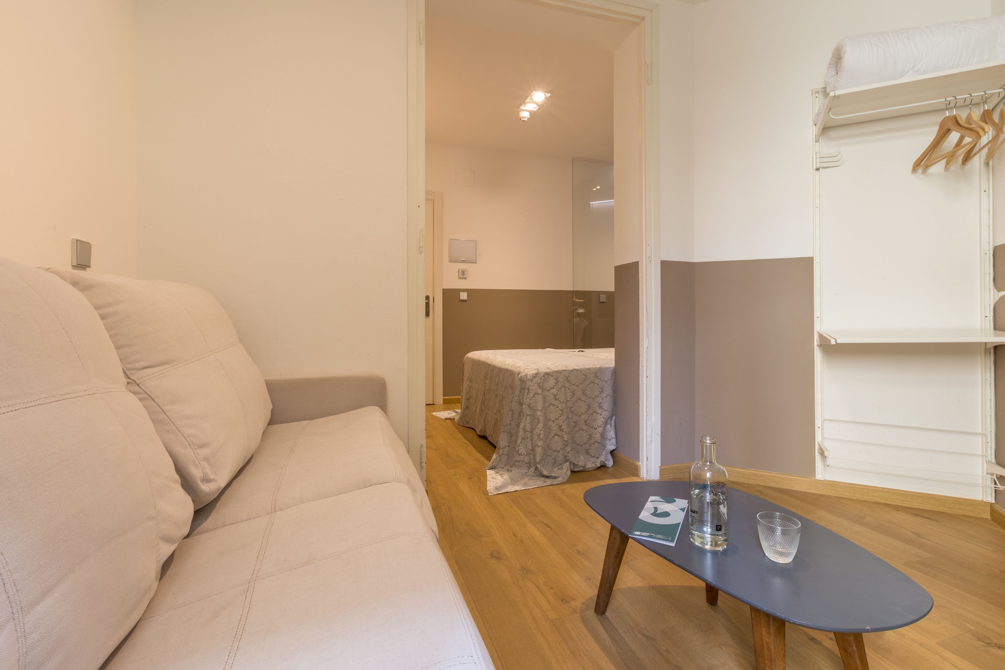 A room with a beige sofa, a small table with a bottle and glass, a bed with a patterned bedspread, and a clothing rack.