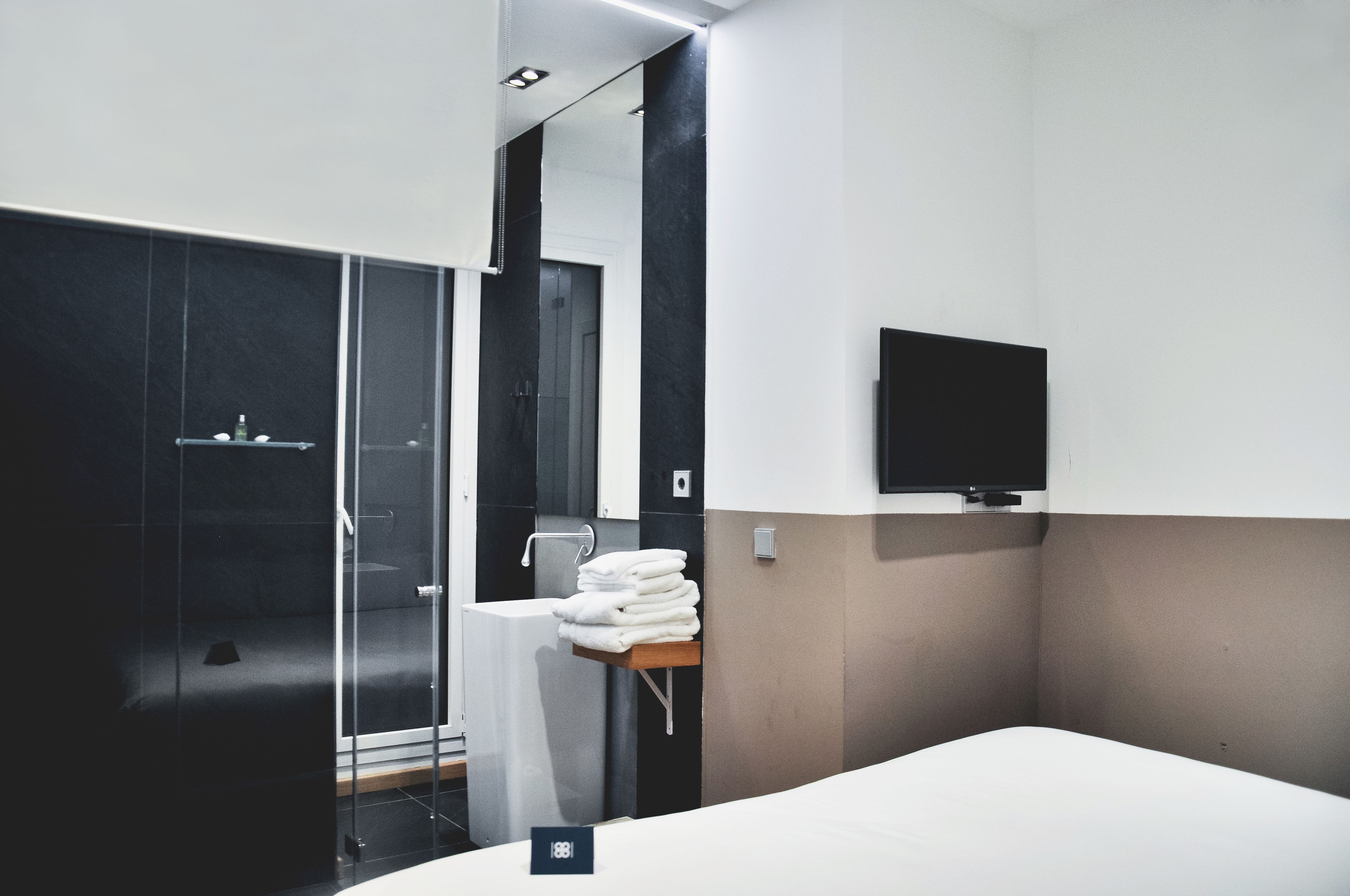 Una habitación de hotel moderna con cabina de ducha de vidrio, una cama, un televisor montado y una pila de toallas blancas sobre una mesita.