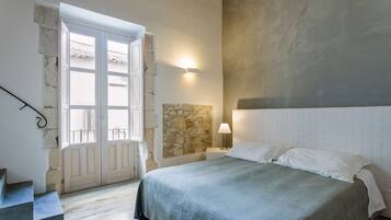 Een slaapkamer met een bed, een stenen muur, een nachtkastje met een lamp en een raam met uitzicht.