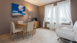 A modern living room with a dining area, a flat-screen TV, and a large window with white curtains. - Garden House   & Eastpark Apartments (Munich)