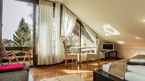 A modern bedroom with a large bed, a desk, and a chair. There is a sliding glass door leading to a balcony with a view of trees. - Garden House   & Eastpark Apartments (Munich)
