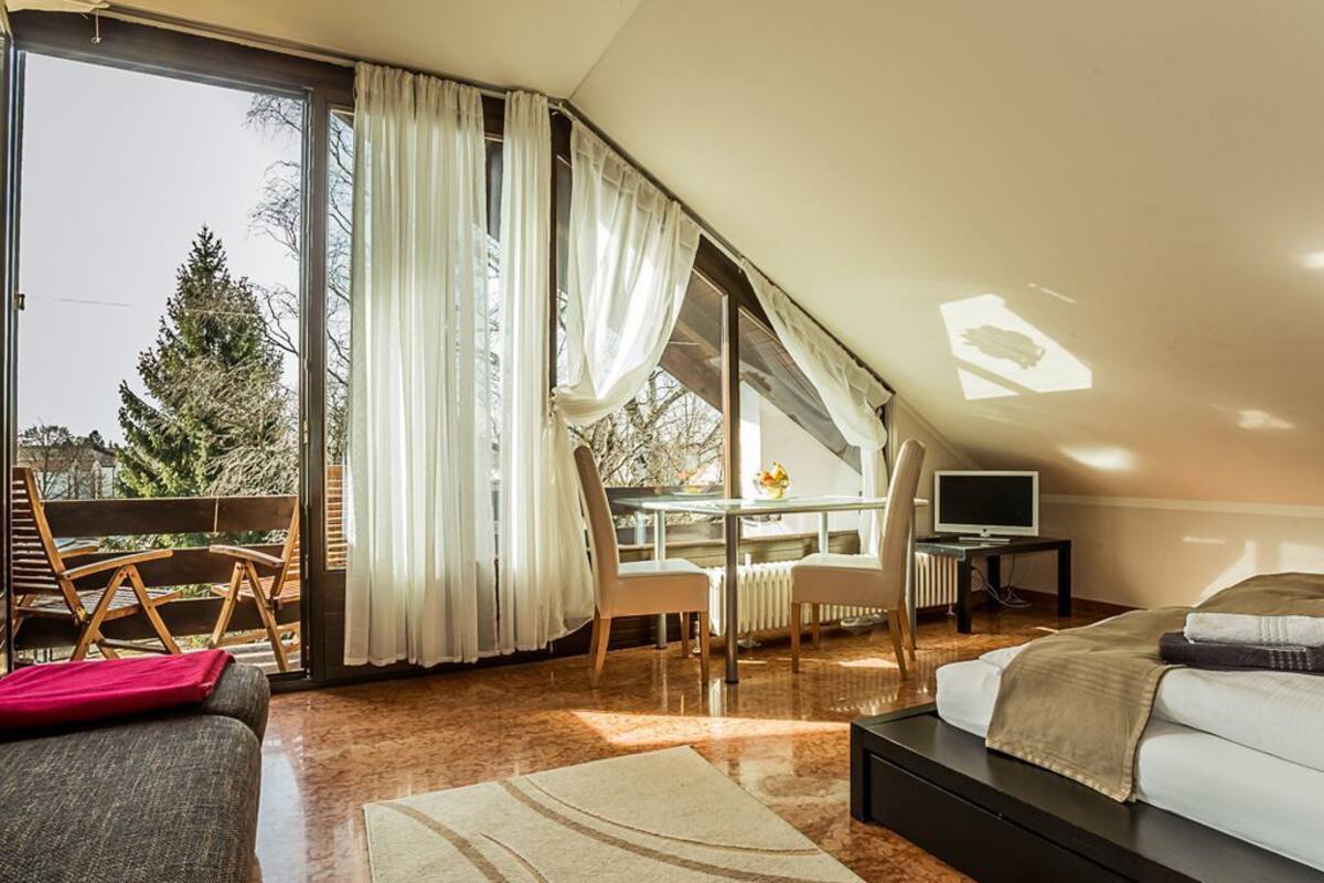 Een moderne slaapkamer met een groot bed, een bureau en een stoel. Er is een glazen schuifdeur die leidt naar een balkon met uitzicht op bomen.