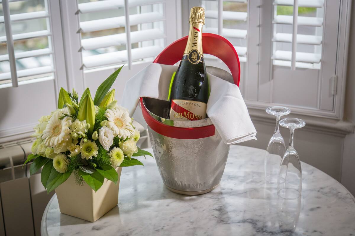 A bottle of champagne in a silver bucket with a red handle, placed on a marble table next to a bouquet of white flowers in a beige vase.