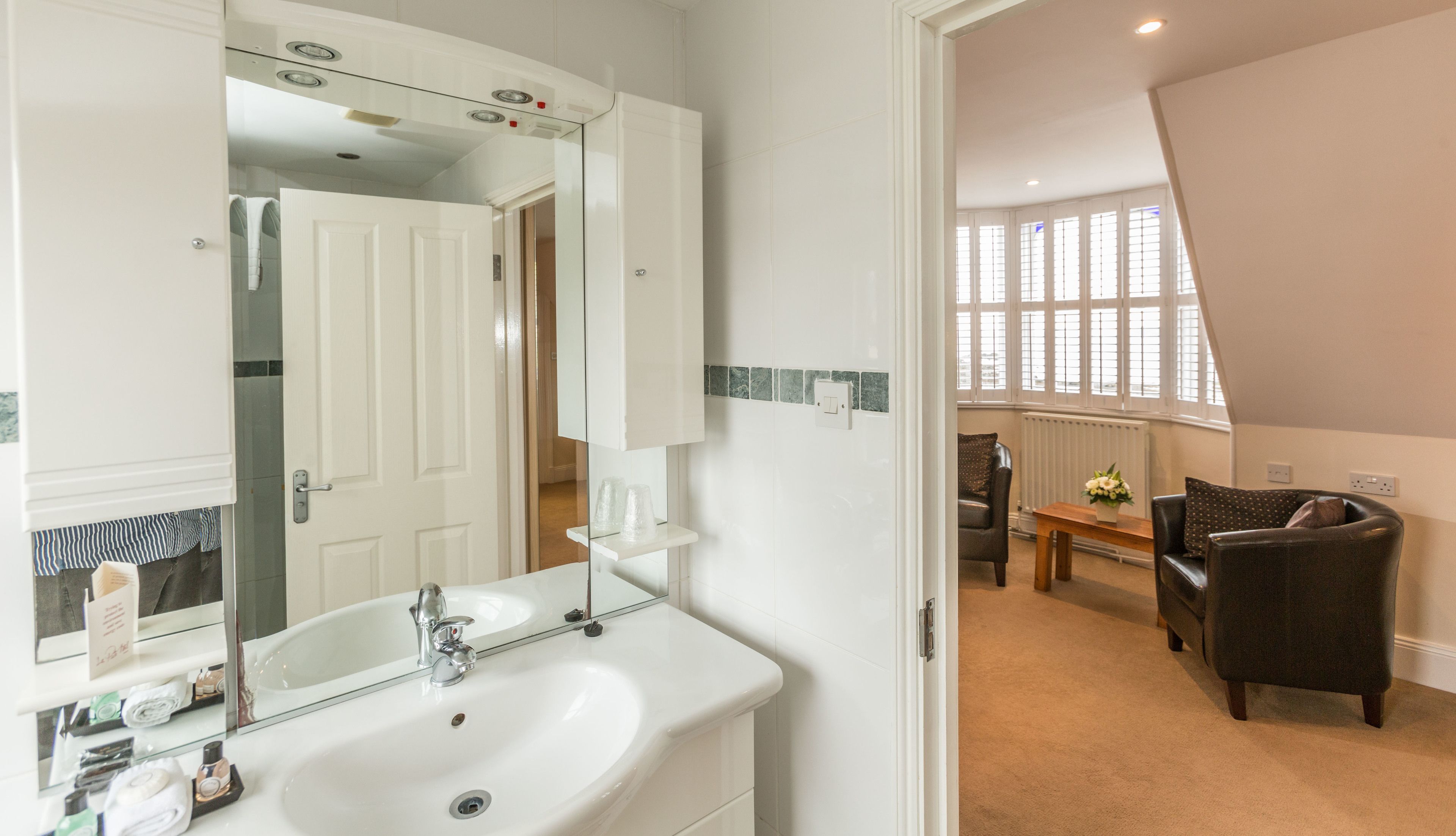 Un baño con un espejo grande, lavamanos blanco y una repisa de vidrio con artículos de tocador. Se ve una puerta que da a una zona de estar con un sofá y una mesa.