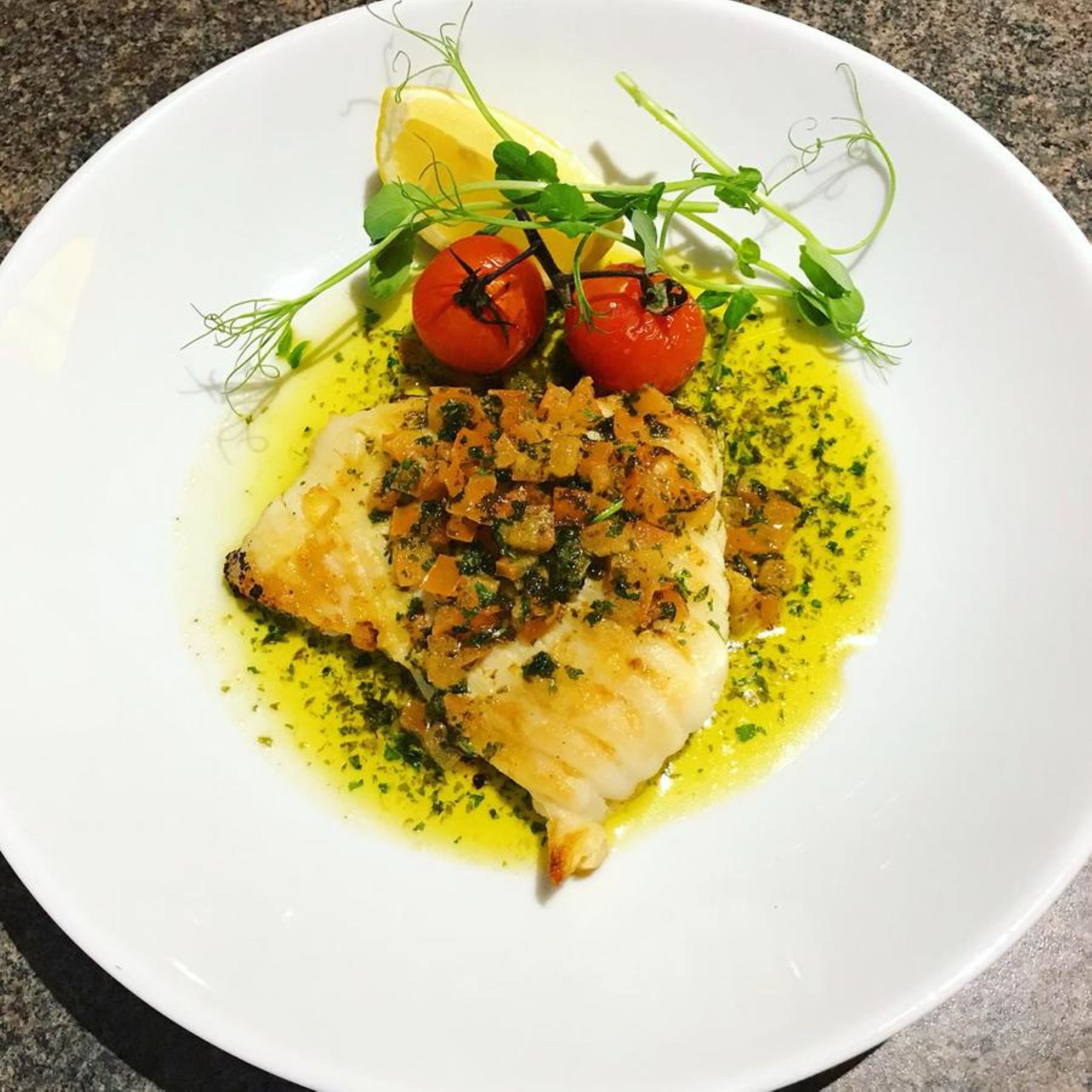 Un plato blanco con un trozo de pescado, tomates cherry y guarnición de hojas verdes.