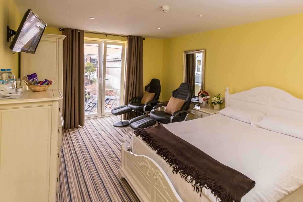 A hotel room with a bed, a desk with a chair, a TV mounted on the wall, and a window with curtains.