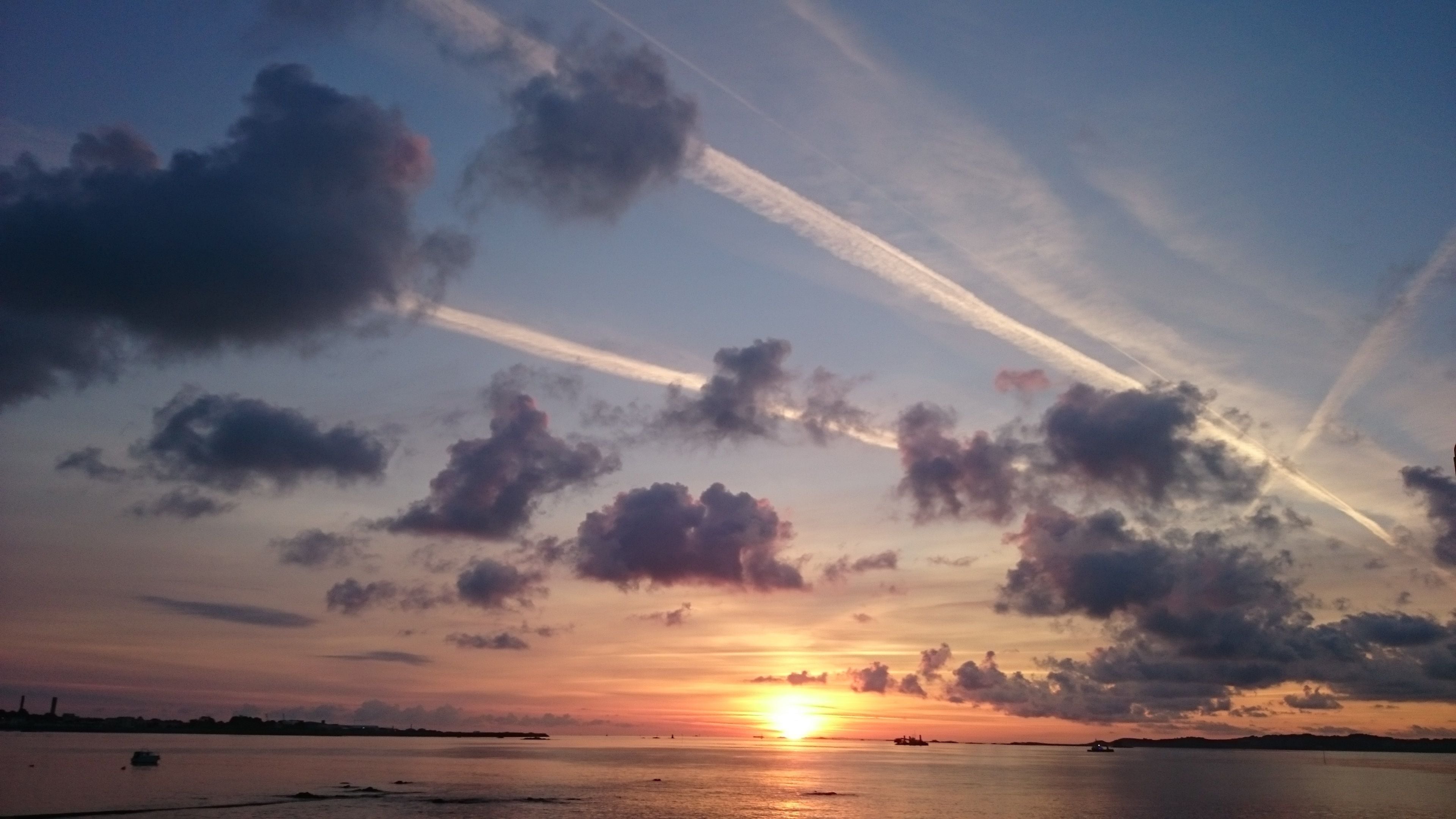 Un coucher de soleil sur une étendue d’eau calme, avec quelques nuages épars et des traînées de condensation.