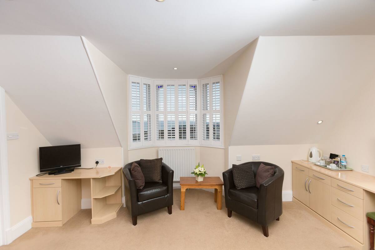A room with a desk, two chairs, a small table with a vase, and a window with blinds.