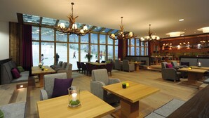 Un espacio interior amplio con mesas y sillas de madera, ventanas grandes que dan a una pista de esquí al aire libre y varios candelabros.