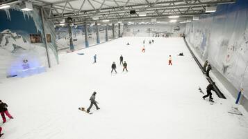 Pista de esqui coberta com neve artificial, teleféricos e um grande mural de uma montanha nevada.