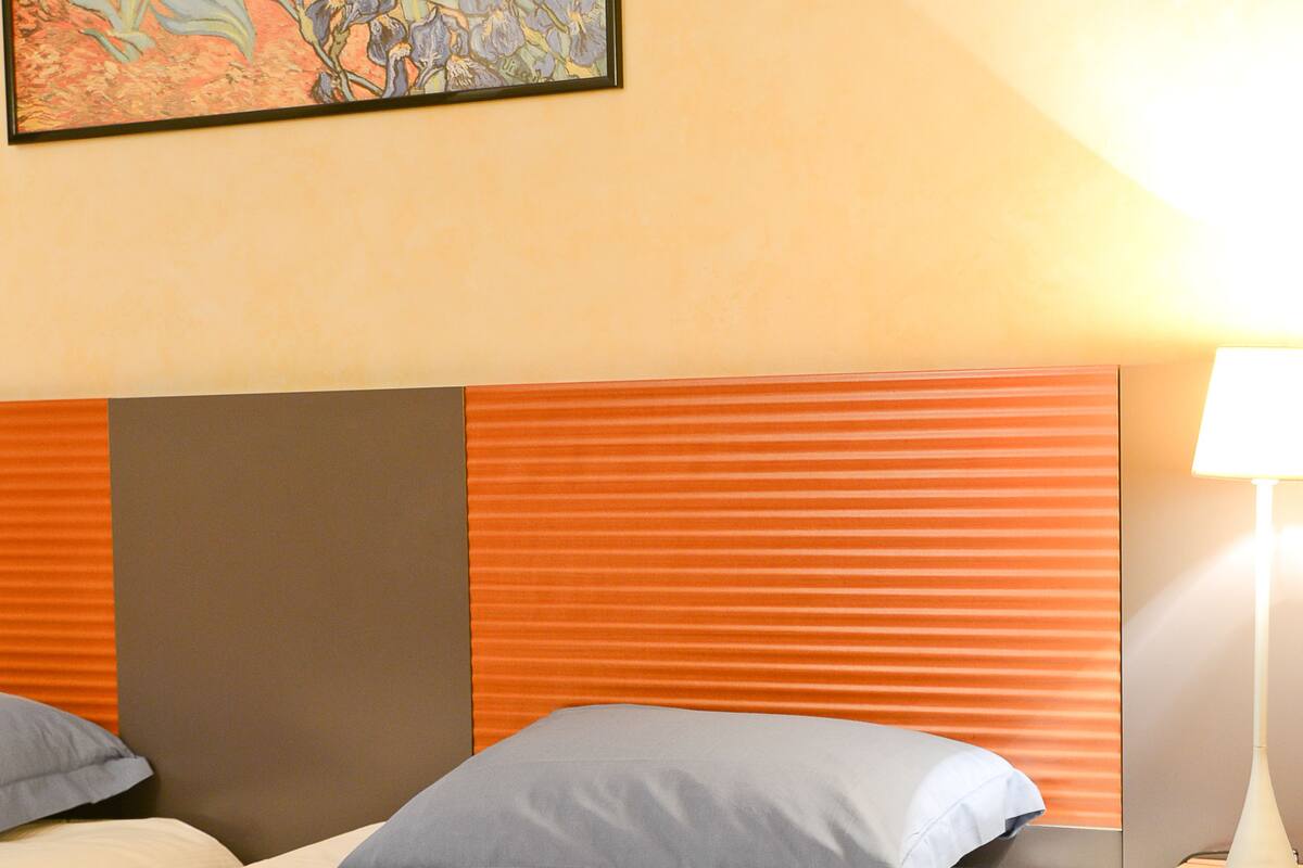 A hotel room with two beds, a headboard with orange and gray panels, a bedside table with a lamp, and a painting on the wall.