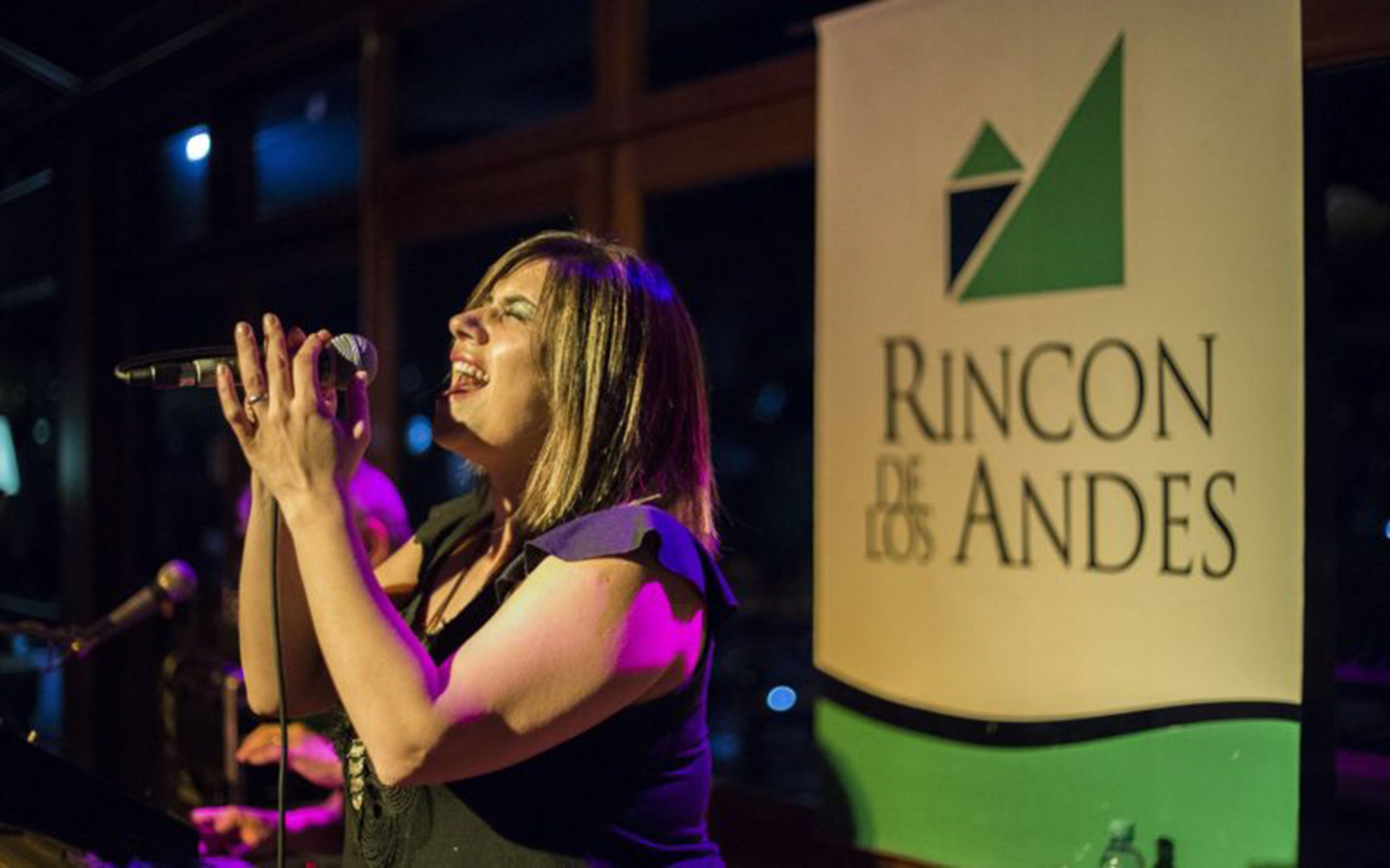 Uma pessoa cantando em um microfone, com uma faixa exibindo "Rincon de Andres" ao fundo.