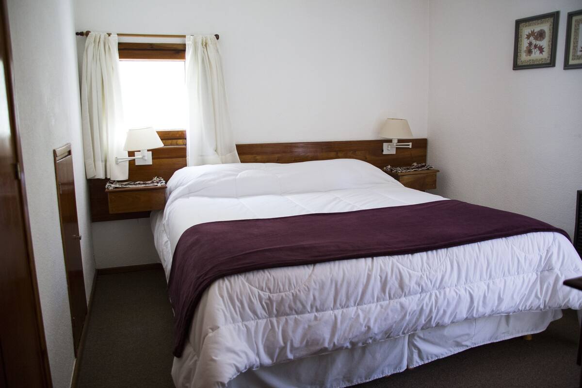 A bedroom with a bed, a nightstand with a lamp, a window with curtains, and a wall with framed pictures.
