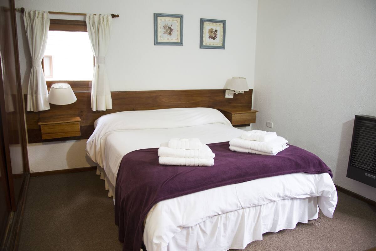 A neatly made bed with white and purple bedding, a wooden headboard, and two wall-mounted lamps.