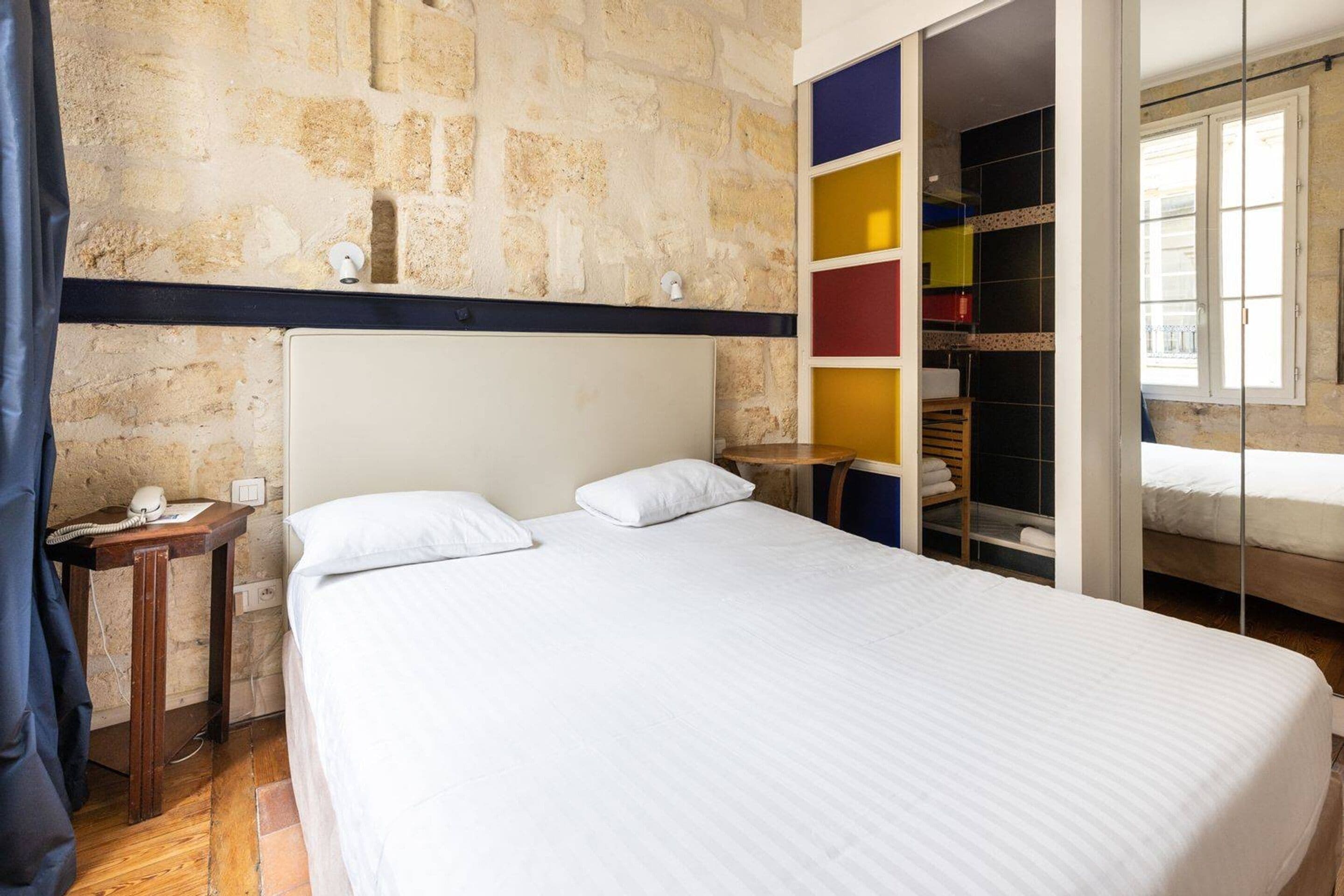 A bedroom with a stone wall, a bed with white bedding, a bedside table with a phone, and a colorful storage unit.