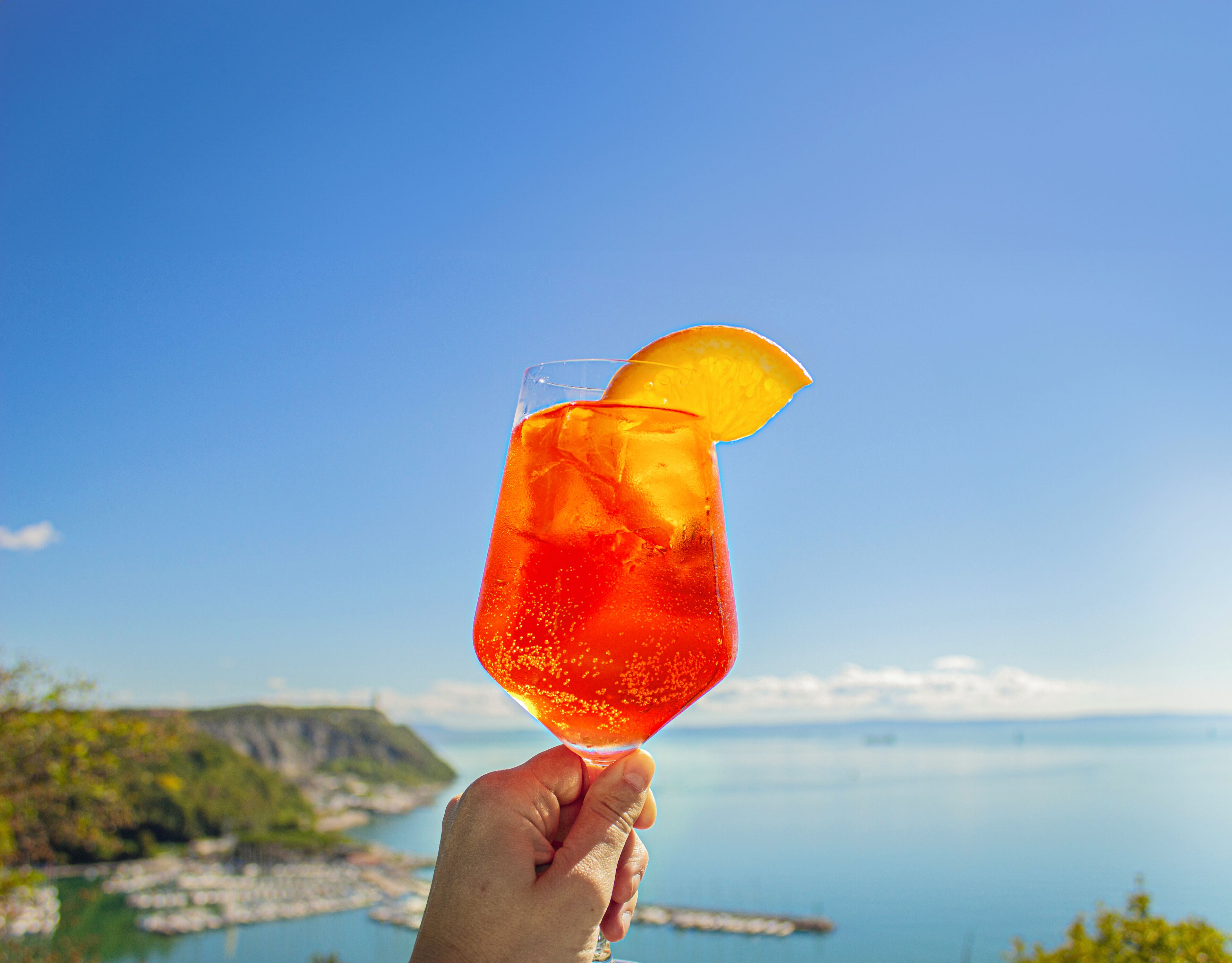 Una mano sostiene un vaso con una bebida roja y una rodaja de naranja, sobre un fondo de cielo azul despejado y un paisaje costero.