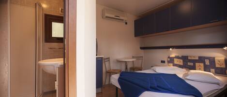 Habitación de hotel pequeña con ducha, una mesita, una cama con manta azul y armarios azul oscuro.