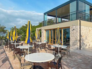 An outdoor patio with round tables and chairs, yellow and blue striped umbrellas, and a stone wall. - Camping Village Mare Pineta Baia Sistiana (Duino-Aurisina)