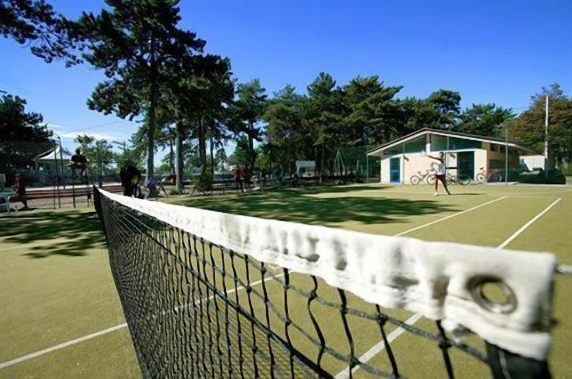 A tennis court with a net in the foreground, surrounded by trees and a building in the background.