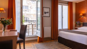 Habitación de hotel con cama, escritorio, silla, ventana con cortinas, radiador y vista a un edificio.
