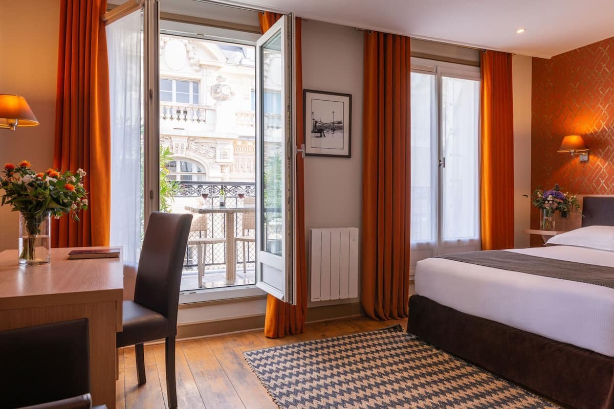 Habitación de hotel con cama, escritorio, silla, ventana con cortinas, radiador y un cuadro en la pared.
