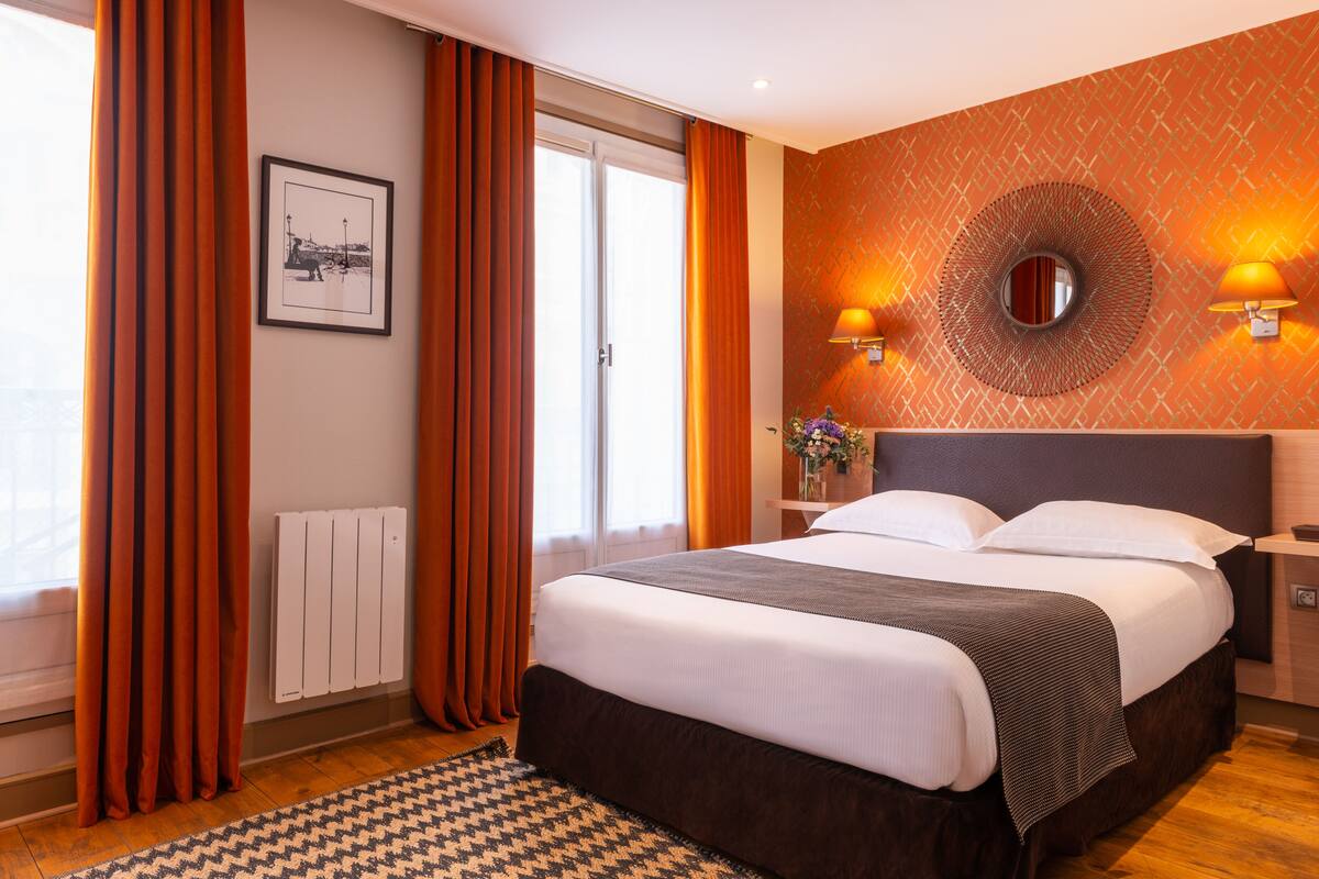 Habitación de hotel con una cama grande, cortinas anaranjadas, un radiador, un cuadro enmarcado y dos lámparas fijadas a la pared.