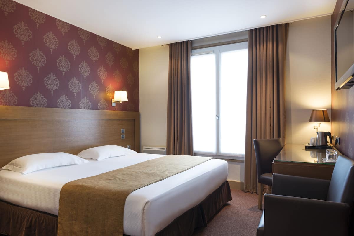 Habitación de hotel con una cama grande, un escritorio con silla, una ventana con cortinas y un papel tapiz estampado en la pared.