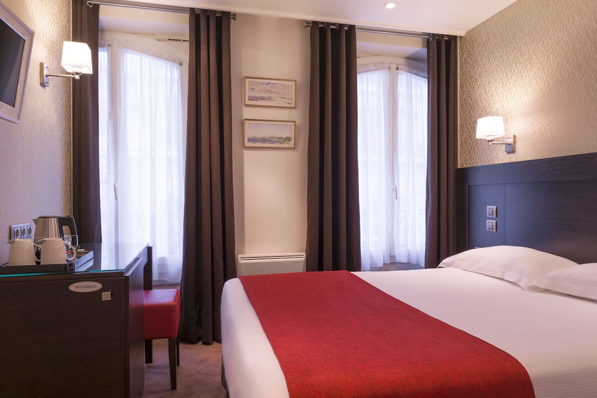 Una habitación de hotel con una cama, un escritorio con minibar y dos ventanas con cortinas.