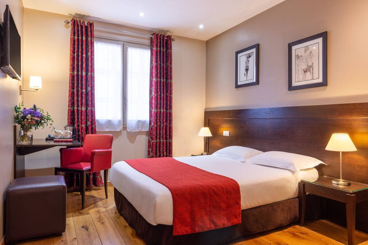 Una habitación de hotel con cama, lámparas de noche, un escritorio con una silla, una mesita con un florero y dos cuadros en la pared.