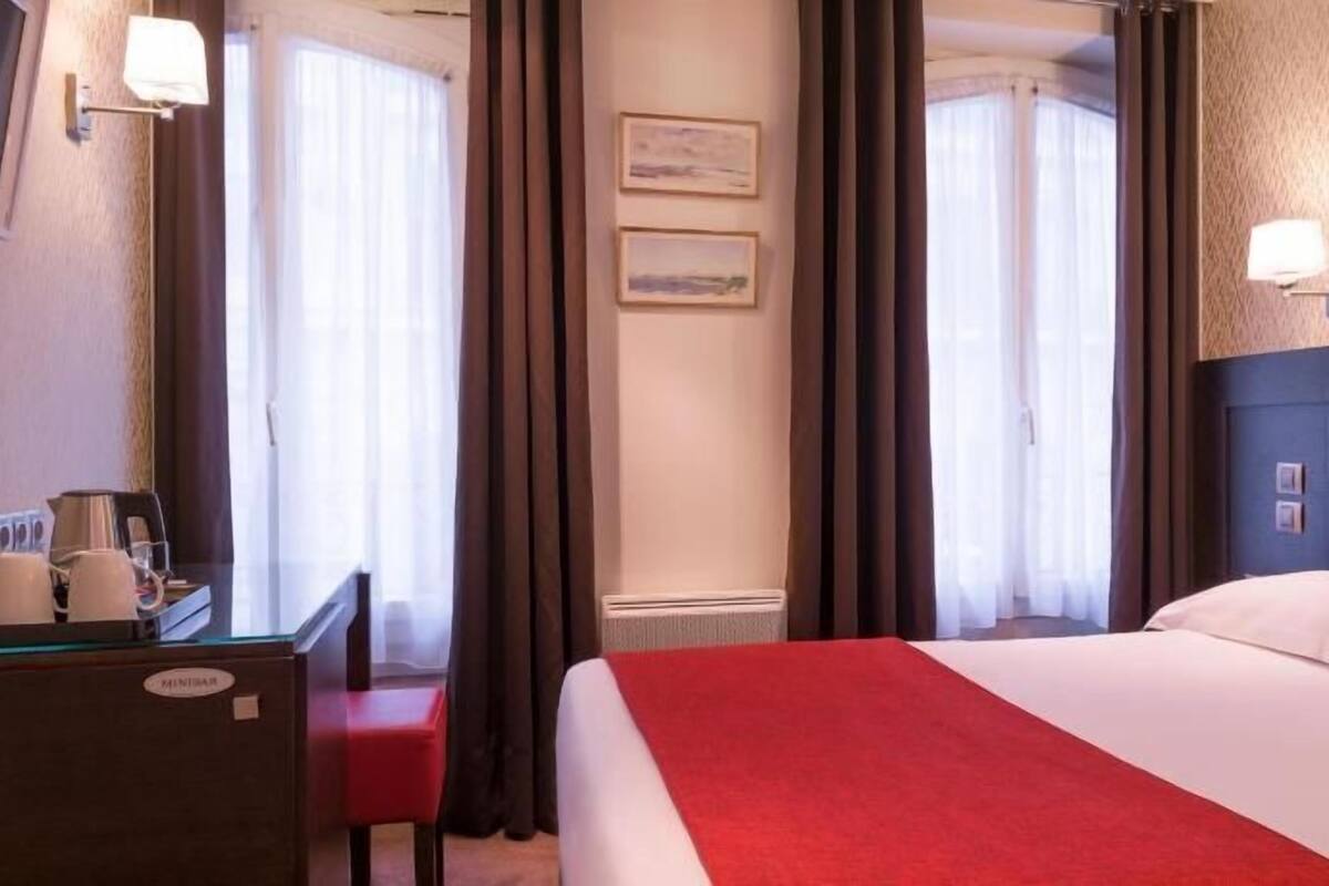 Una habitación de hotel con una cama, un escritorio con cafetera, una silla y cortinas.